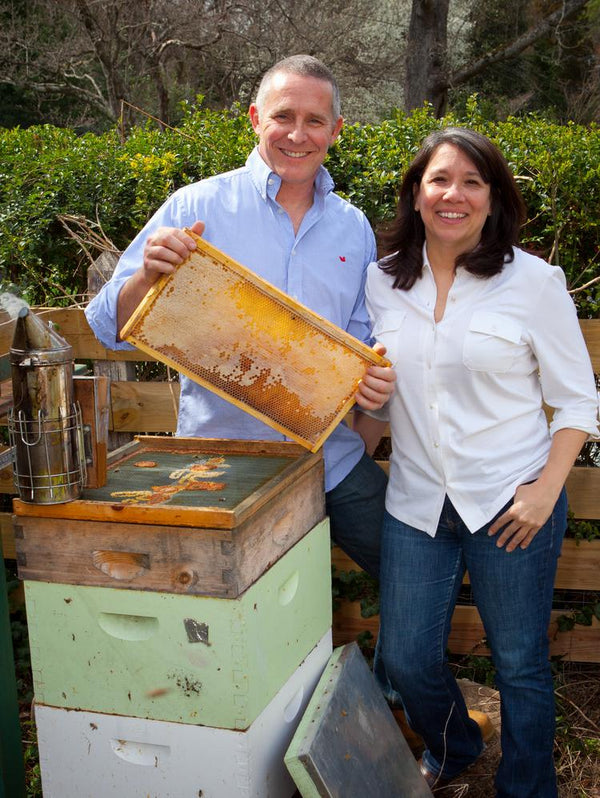 Cloister Honey founders Randall and Joanne smiling beside a stack of beehives, Randall holding up a frame of capped honeycomb with a smoker on top of the boxes.