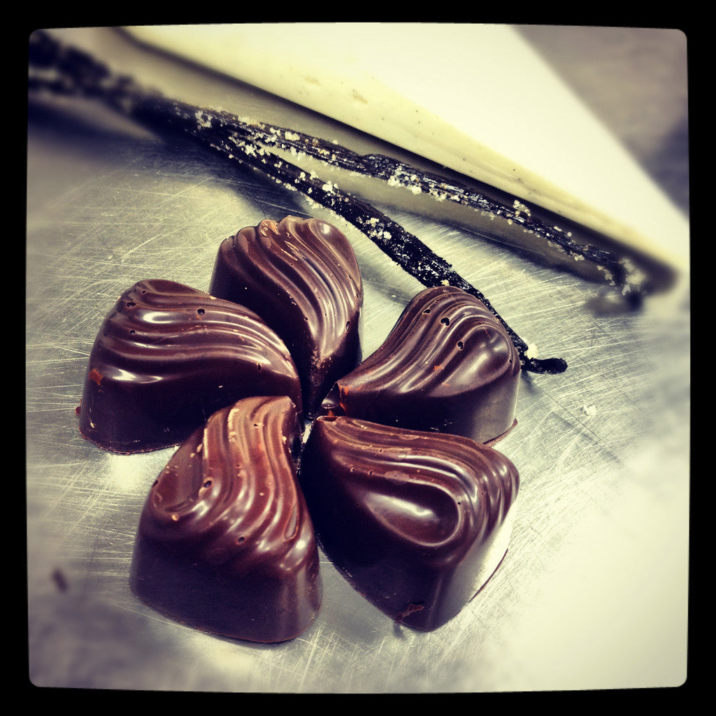 Five Vanilla Bean Bon Bons arranged in a circle around vanilla bean pods, the dark chocolate shells in a fanned-shell shape. Main image
